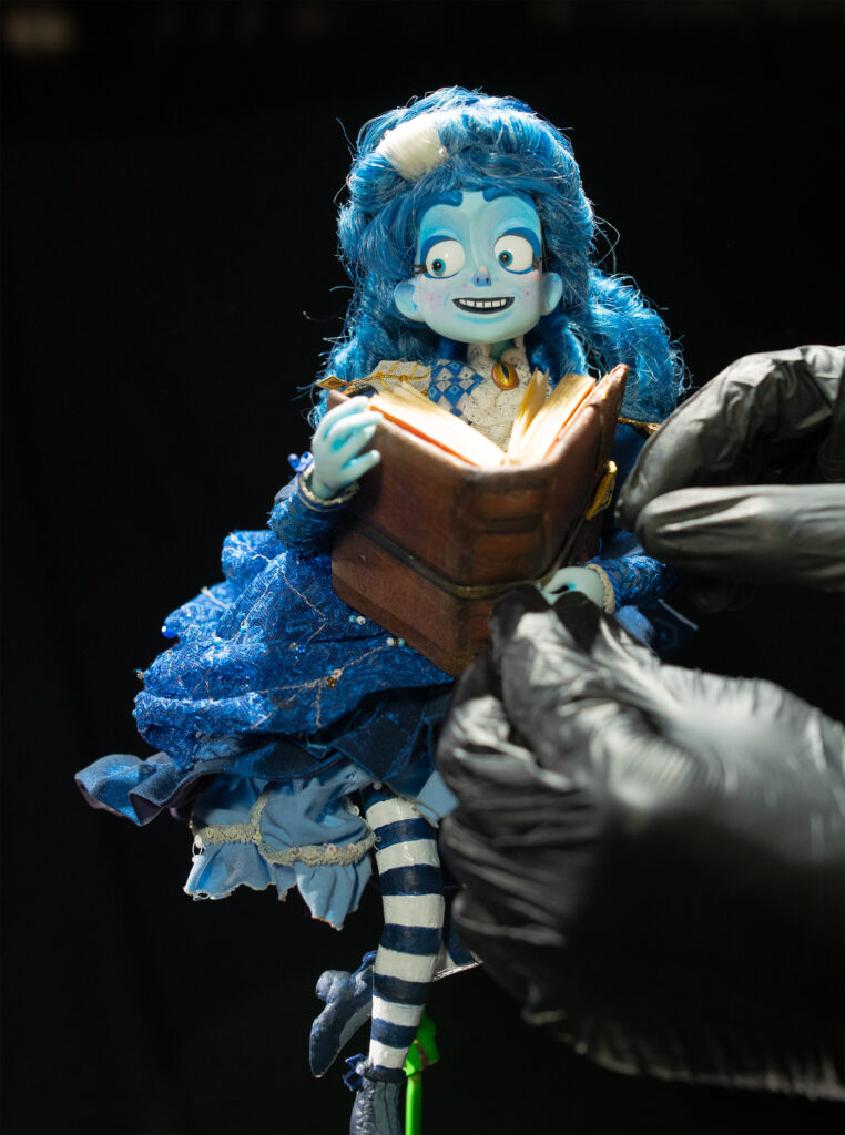 Manos con guantes negros ajustando una marioneta de Frankelda de cabello azul que sostiene un libro, durante el proceso de animación stop-motion.