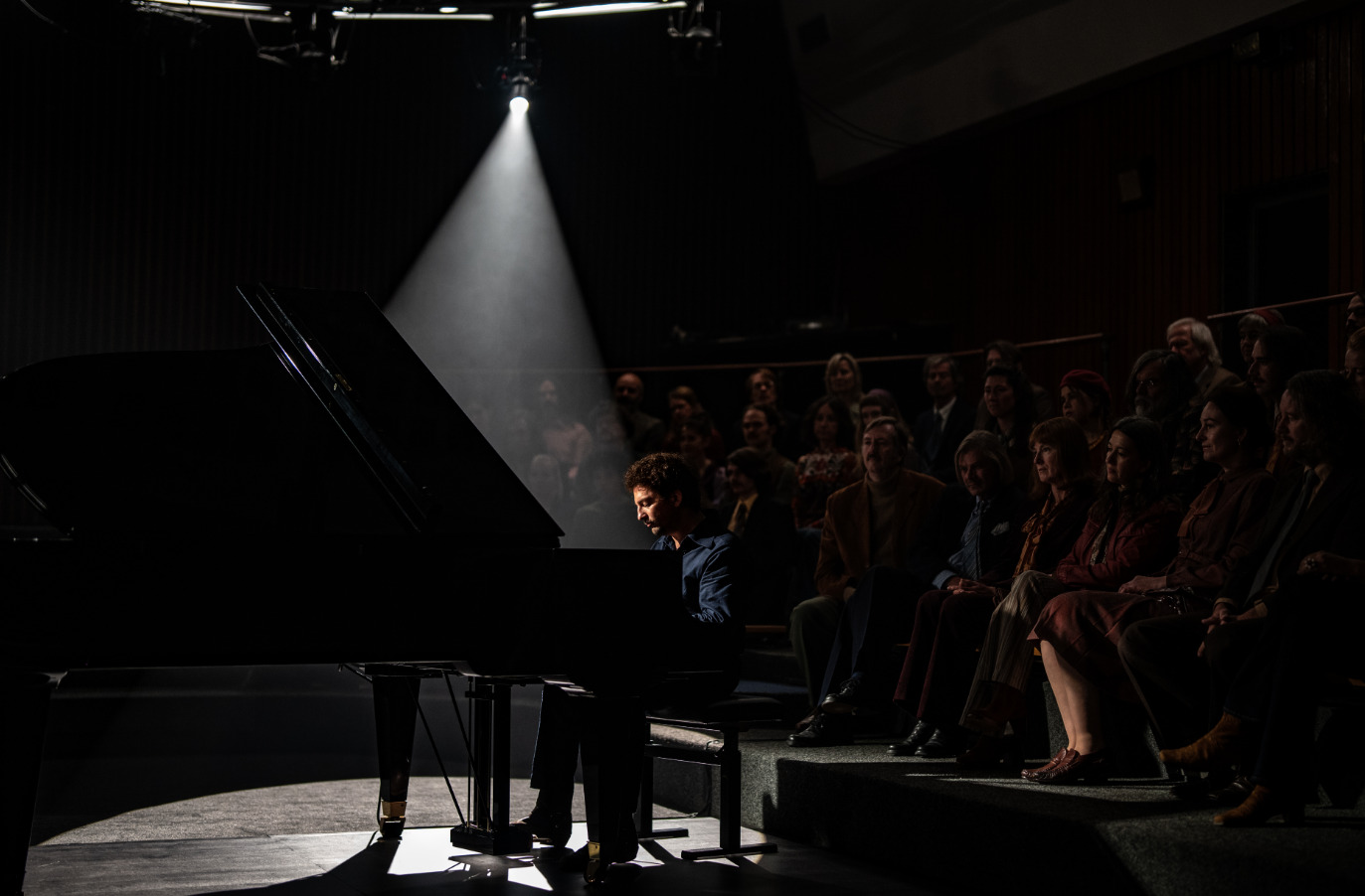 Fotograma de la película "Köln 75", con el actor John Magaro como Keith Jarrett tocando un piano de cola en un escenario, con público en la oscuridad.