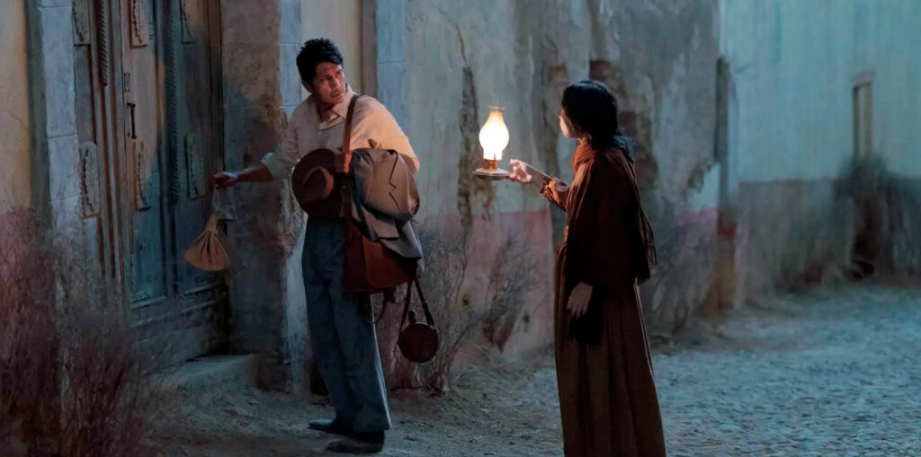 Fotograma de la película "Pedro Páramo", con dos personajes en una calle oscura, una mujer sosteniendo una lámpara y un hombre llegando a una puerta.