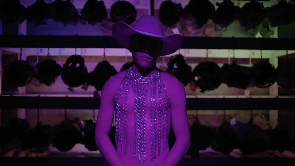 Fotograma de la película "En el camino", de David Pablos, mostrando a un hombre con sombrero vaquero y camiseta de brillos bajo luz violeta.