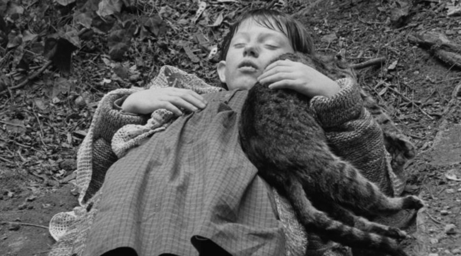 Fotograma en blanco y negro de "Sátántangó", mostrando a una niña abrazando un gato, recostada en el suelo.