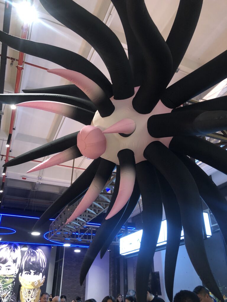 Escultura inflable 'Electric Bloom x' de la artista visual Laura Elena Garduño, en exposición durante el evento 'Entre raíces y ciudad' en Level Campestre León. La pieza cuelga del techo y presenta tentáculos negros que contrastan con un centro blanco y protuberancias rosadas.