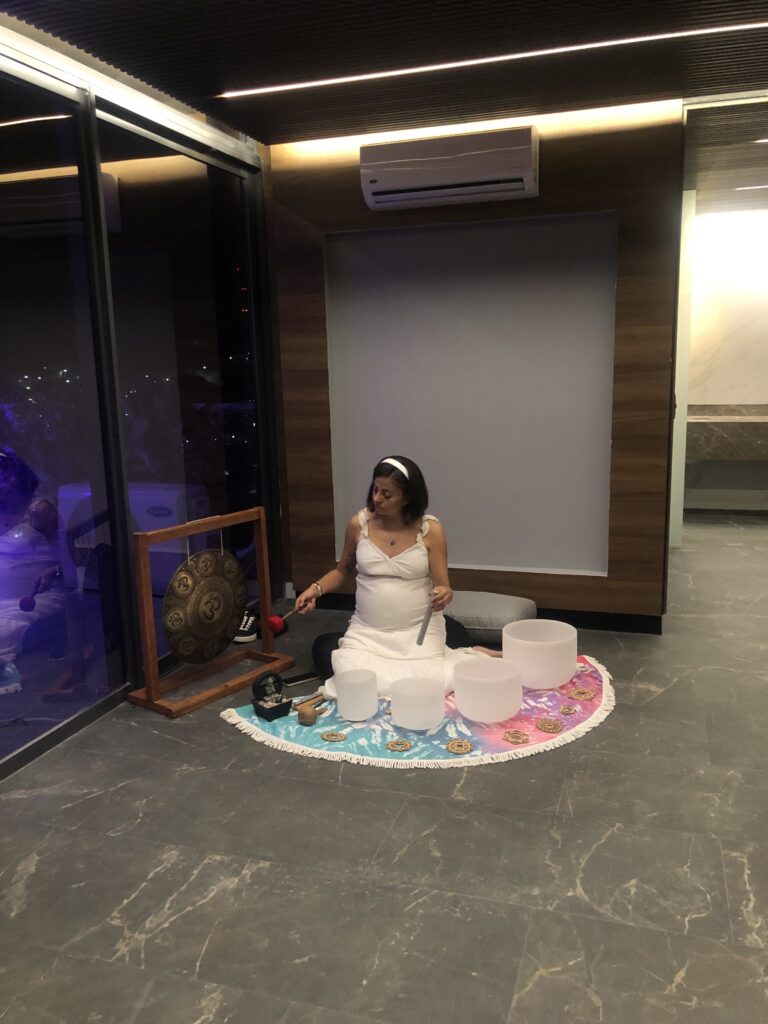 Sonoterapeuta realizando una sesión de sonoterapia con cuencos tibetanos de cuarzo y un gong, como parte del componente sensorial durante el recorrido de inauguración de las amenidades en Adamant Grand León, con vistas nocturnas de la ciudad.