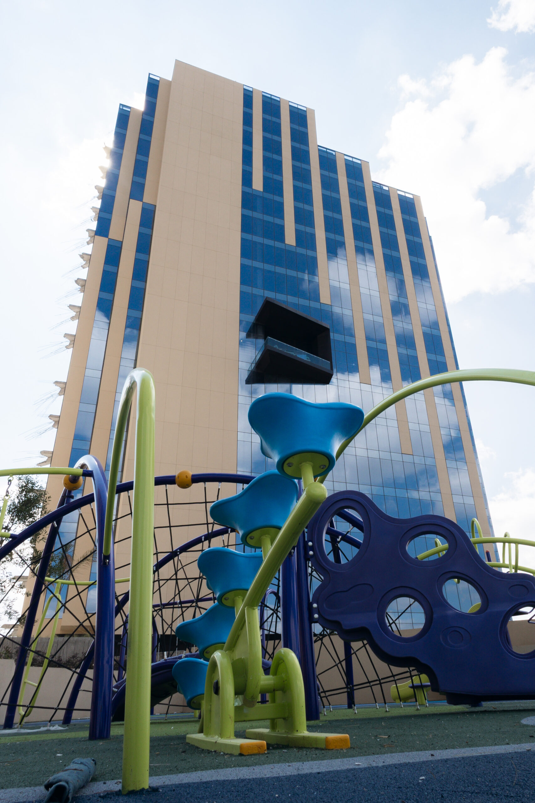 Vista exterior de la moderna fachada del edificio Adamant Grand León, con paneles beige y cristal azul reflejando el cielo, y un balcón negro sobresaliente. En primer plano, se ve un área de juegos infantiles. Desarrollo de Milk Life Investments.