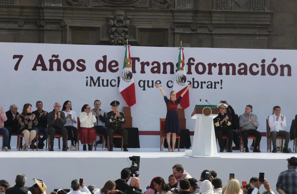 La presidenta Claudia Sheinbaum levanta los brazos en señal de triunfo en el Zócalo, celebrando frente al banner "7 Años de Transformación".