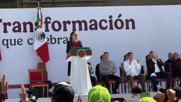 Claudia Sheinbaum dando un discurso en el Zócalo de la Ciudad de México, celebrando los siete años de la Cuarta Transformación, con la bandera de México detrás.