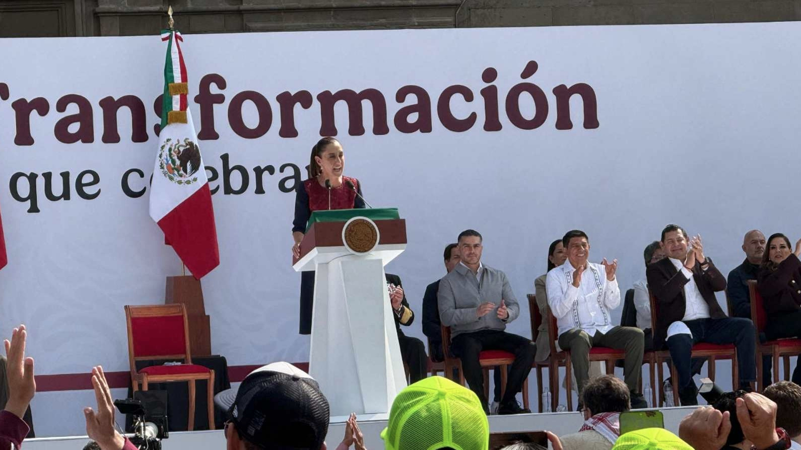 Claudia Sheinbaum dando un discurso en el Zócalo de la Ciudad de México, celebrando los siete años de la Cuarta Transformación, con la bandera de México detrás.