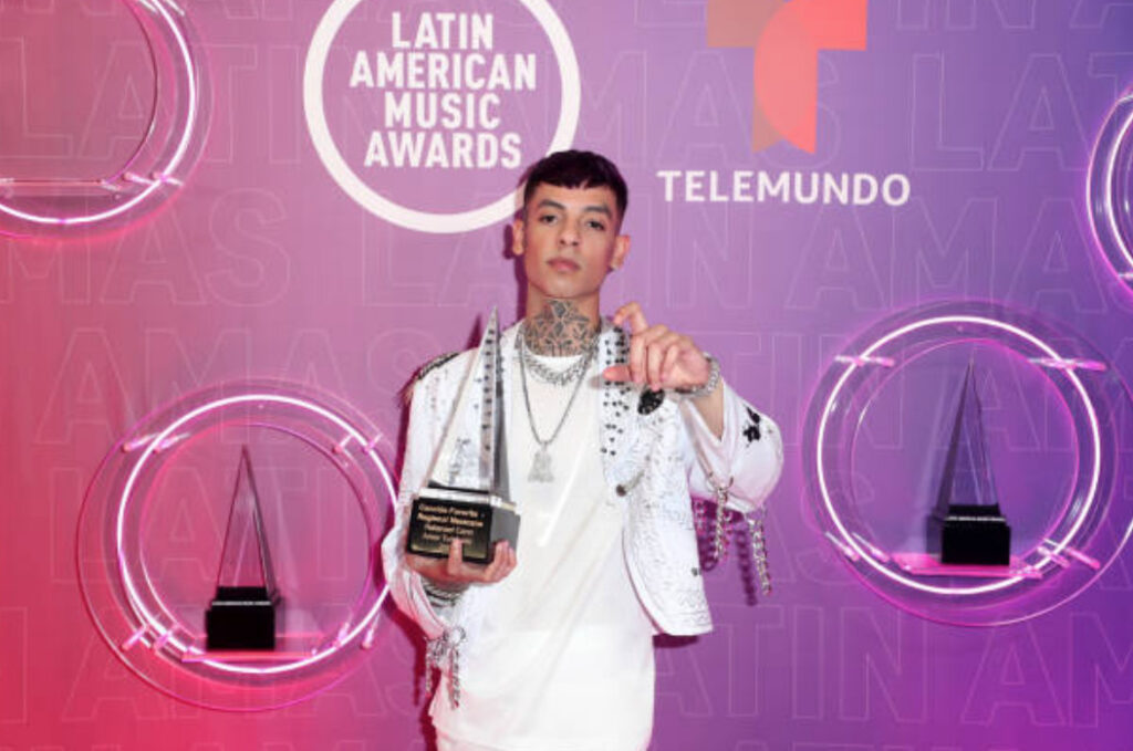 Natanael Cano sonriendo y sosteniendo su premio transparente de 'Canción Favorita - Regional Mexicano' por 'Amor Tumbado' en los Latin American Music Awards 2021, vistiendo una chaqueta blanca decorada. Fondo rosa con logos de los Latin AMAs y Telemundo.