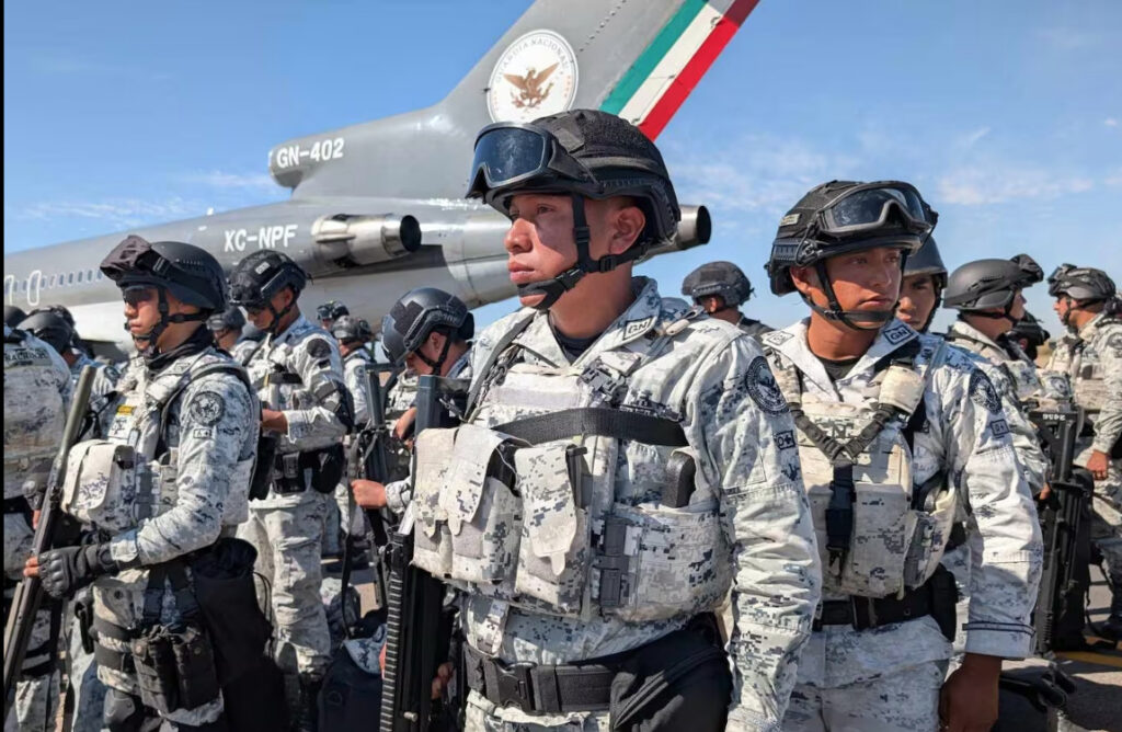 Un grupo de soldados de la Guardia Nacional de México con uniforme de camuflaje pixelado y equipo táctico completo están de pie en formación en la pista de un aeropuerto, frente a un gran avión de transporte militar gris con la bandera de México y el logo de la Guardia Nacional en el estabilizador vertical. Foto: Julián Ortega García.
