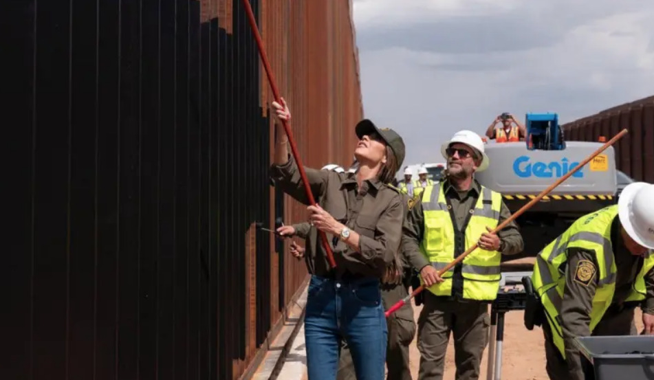 Kristi Noem, exsecretaria de Seguridad Nacional, usando un rodillo de mango largo para pintar de negro las barras del muro fronterizo entre Estados Unidos y México.