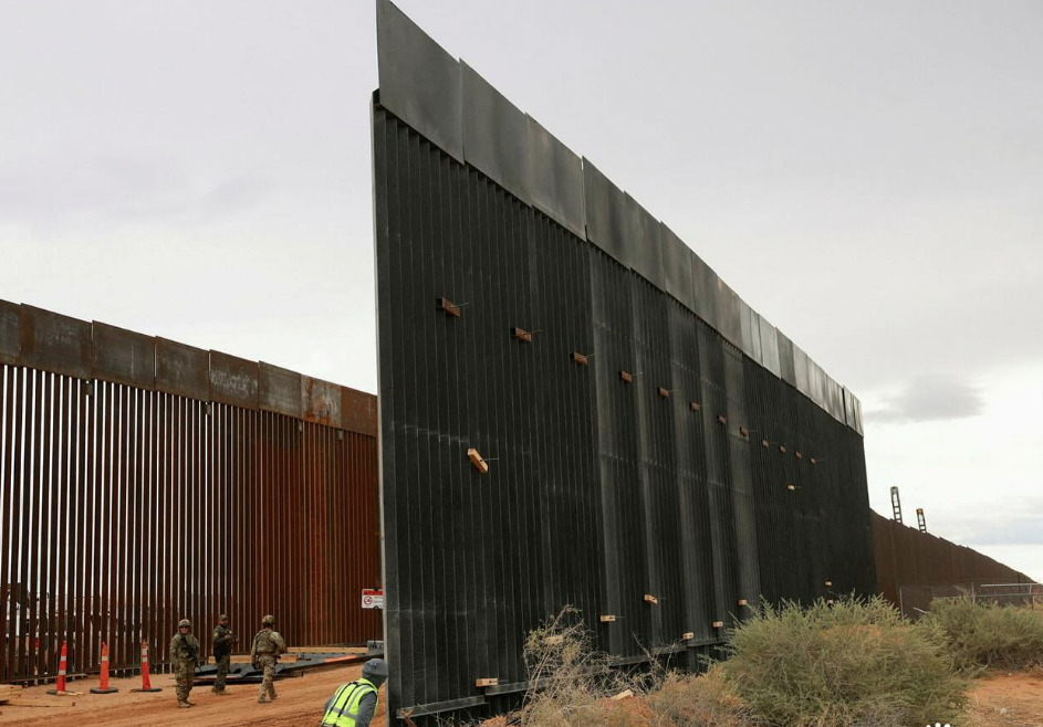 Perspectiva lateral de una sección terminada del muro fronterizo negro de gran altura, con personal de seguridad y trabajadores en la base de la estructura en una zona desértica.