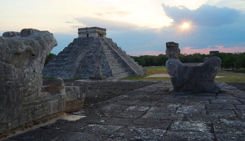 Primer plano de escultura Maya con la Pirámide de El Castillo al fondo en Chichén Itzá, Yucatán, bajo un atardecer nublado.