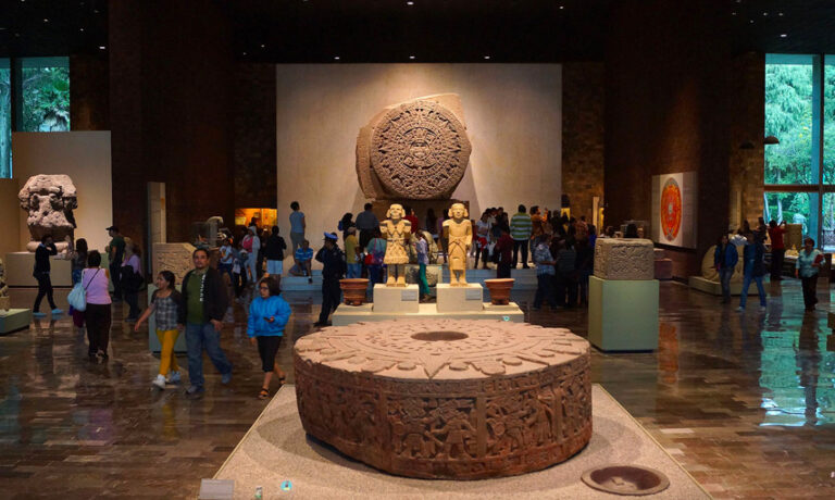 Vista interior de la Sala Mexica del Museo Nacional de Antropología con turistas admirando la Piedra del Sol y otras esculturas aztecas monumentales.
