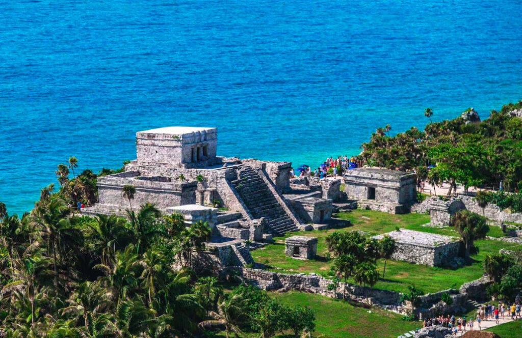 Vista aérea de El Castillo y templos mayas en la zona arqueológica de Tulum frente al mar Caribe turquesa con turistas recorriendo el sitio.