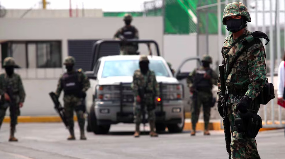 Soldado de la Armada de México con equipo táctico y fusil de asalto vigilando una calle en Mazatlán, con otros efectivos y un vehículo oficial al fondo.