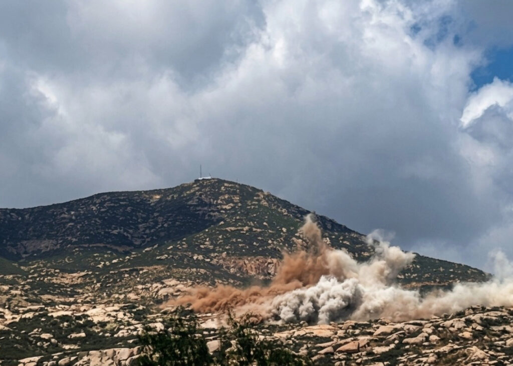 Gran nube de humo marrón y blanco elevándose tras una detonación en la ladera rocosa y cubierta de matorrales del Cerro Cuchumá, cerca de la frontera México-EE. UU. En la cima se observa una torre de comunicaciones.