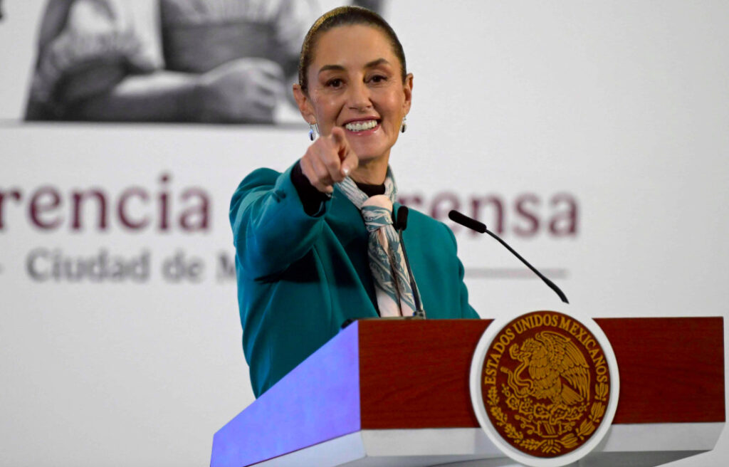 La presidenta Claudia Sheinbaum sonriendo en un podio con el escudo nacional de México, señalando hacia el frente con el dedo índice durante una conferencia de prensa.