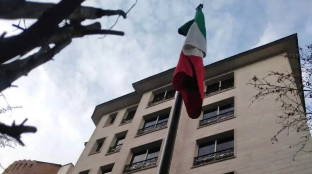 Retrato en plano general de la fachada de un edificio moderno con ventanas balconadas, donde cuelga verticalmente la bandera de México (verde, blanco y rojo). Fondo de cielo nublado y ramas oscuras en primer plano.