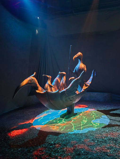 Escultura de bronce de Leonora Carrington que representa un barco con forma de pájaro transportando a otras figuras de aves estilizadas, iluminada por una proyección de agua azul y verde en la base.