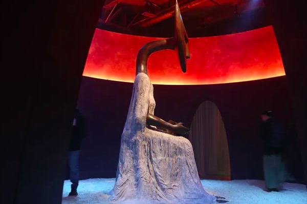 Escultura de figura femenina con cabeza alargada y estilizada dentro de una sala circular iluminada con tonos rojos en la exposición Laberinto Mágico.