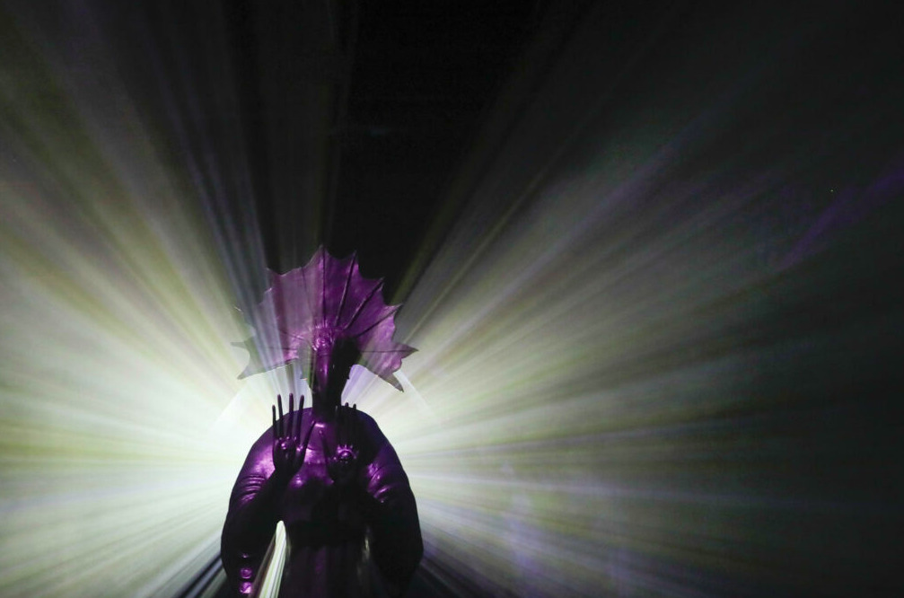 Escultura de bronce de un personaje surrealista de Leonora Carrington con túnica y rostro alargado, rodeado de proyecciones circulares de luz dorada y azul en una sala inmersiva.