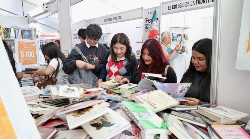 Grupo de personas caminando entre stands de libros bajo carpas blancas en la FIL UABC, con señalética de la Universidad Autónoma de Baja California al fondo.