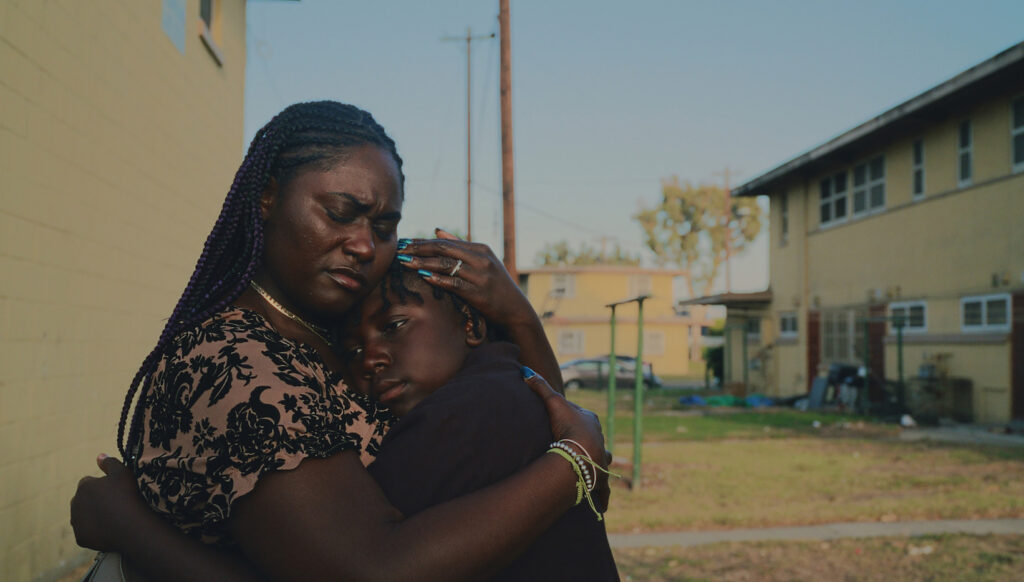 Fotograma de la película 'If I Go Will They Miss Me' que muestra a una mujer con trenzas moradas abrazando protectoramente a un niño en un entorno residencial exterior.