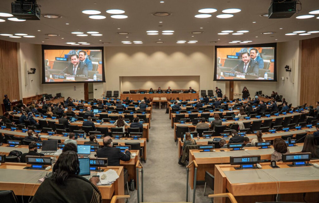 Vista panorámica de una sala de conferencias de la ONU en Nueva York, llena de delegados y con pantallas gigantes que muestran a Ismael Burgueño Ruiz, alcalde de Tijuana, hablando durante una sesión con el Comité de Expertos en Administración Pública.