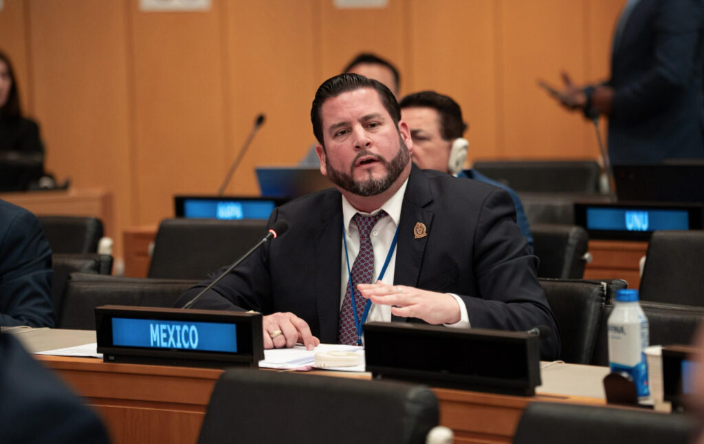 Ismael Burgueño Ruiz, presidente municipal de Tijuana, sentado tras un letrero que dice "MEXICO" en una sala de conferencias de la ONU, hablando con ademanes durante una sesión del Comité de Expertos en Administración Pública.