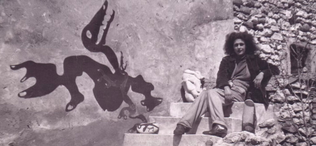 Fotografía antigua en blanco y negro de Leonora Carrington sentada en unas escaleras exteriores junto a una pintura mural de una criatura fantástica.