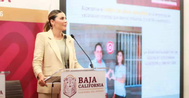 La Gobernadora Marina del Pilar Ávila Olmeda de pie en un podio con el escudo de Baja California, hablando durante una conferencia de prensa ante una pantalla de presentación que muestra un informe sobre salud.