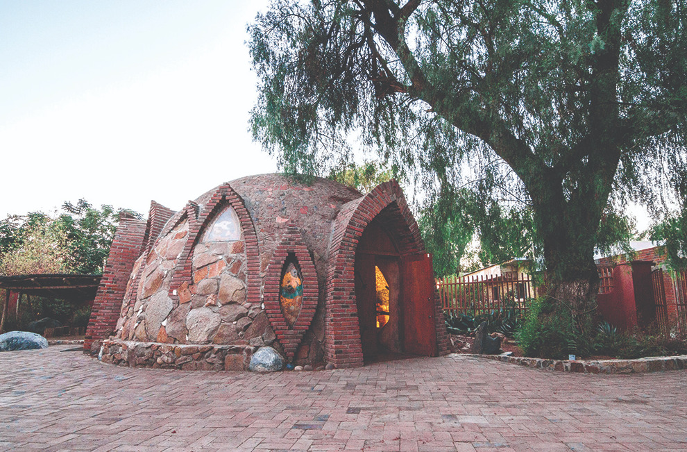 Estructura circular de piedra y ladrillo con techo de domo que representa una vivienda tradicional Kumiai, situada en un patio empedrado bajo la sombra de un gran árbol en el Museo Comunitario de Tecate.