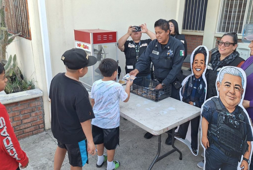 Oficial de la Policía Municipal de Tijuana entregando refrigerios a niños durante un evento de proximidad social del programa Cine en tu Comunidad, junto a figuras de cartón de agentes municipales.