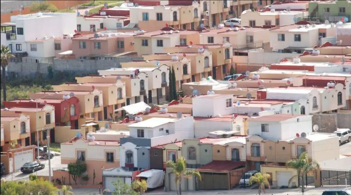 Vista aérea de un fraccionamiento de vivienda social con casas uniformes de dos niveles en tonos terracota y crema en Baja California.