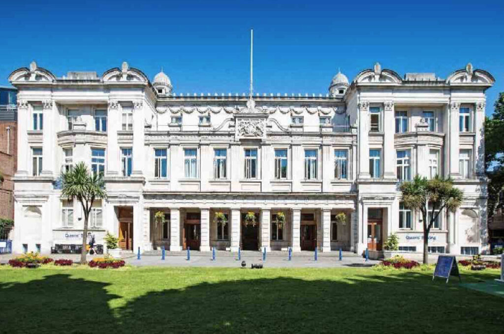 Fachada neoclásica del edificio Queens' Building de la Queen Mary University of London frente a un jardín, institución vinculada al conflicto de becas del IPN.
