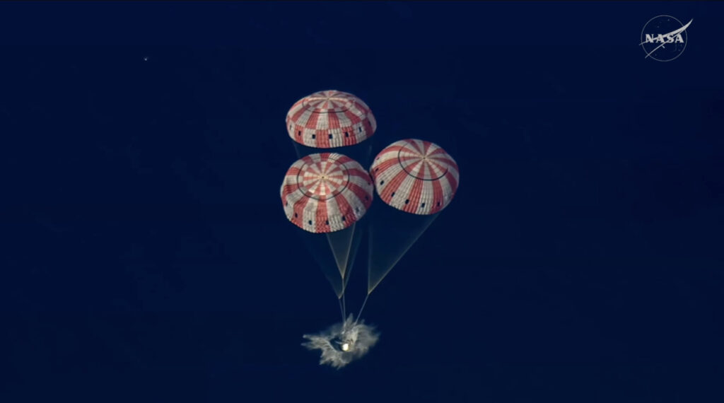 Vista cenital de la cápsula Orión impactando el agua azul profundo del Océano Pacífico, con sus tres paracaídas de franjas rojas y blancas completamente desplegados sobre ella. Crédito: NASA.