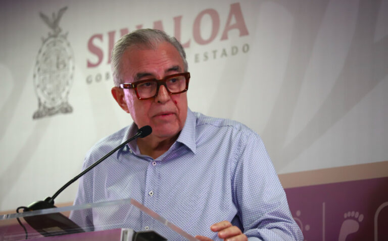 Retrato del gobernador de Sinaloa, Rubén Rocha Moya, vistiendo gafas de montura marrón y una camisa azul claro de puntos, hablando frente a un podio con el logotipo del Gobierno de Sinaloa de fondo.