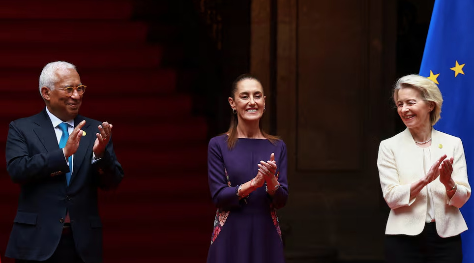 El presidente del Consejo Europeo, António Costa, la presidenta de México, Claudia Sheinbaum, y la presidenta de la Comisión Europea, Ursula von der Leyen, de pie sonriendo y aplaudiendo en Palacio Nacional frente a una bandera de la Unión Europea.