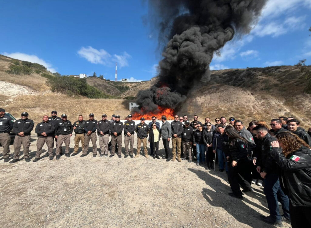 Personal de la Unidad de Auditoría Interna de la FGR y autoridades de seguridad posan frente a la columna de humo de una incineración de narcóticos en un predio abierto bajo el cielo azul.