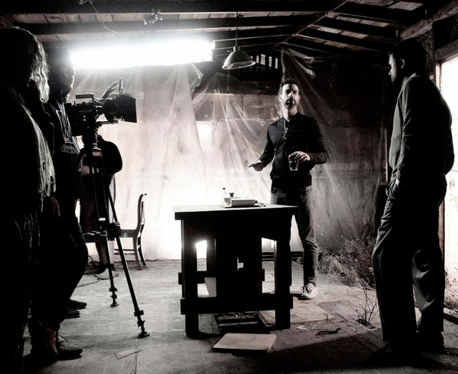 Fotografía en blanco y negro detrás de cámaras del director David Pablos de pie junto a una mesa de madera en un set de filmación rústico, conversando con un actor mientras el equipo técnico prepara la cámara de cine a la izquierda.