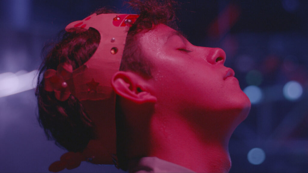 Primer plano de perfil de un joven con los ojos cerrados vistiendo una corona de cartón decorada, iluminado por luces de neón en tonos rojos y púrpuras con luces de fondo desenfocadas.