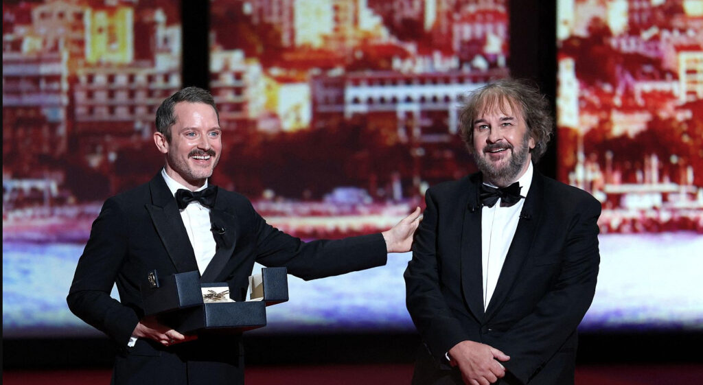 Elijah Wood con esmoquin sostiene el estuche de la Palma de Oro mientras Peter Jackson sonríe a su lado en el escenario del Festival de Cannes.