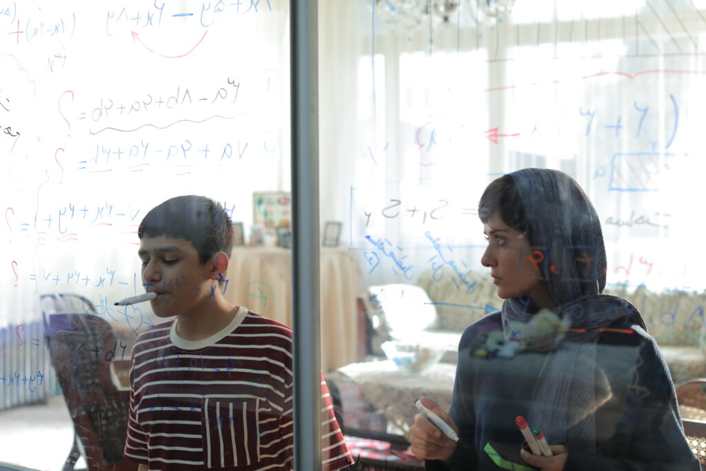 Una mujer con hiyab gris observa fijamente a un joven que escribe ecuaciones matemáticas en un cristal transparente en una escena de la película 'Ella y su hijo'.