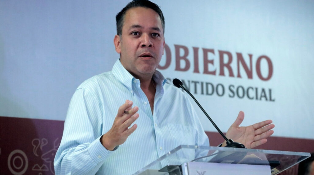 Enrique Díaz Vega con camisa de rayas claras habla en un podio con micrófonos, con el logotipo institucional de Sinaloa "Gobierno con sentido social" al fondo.