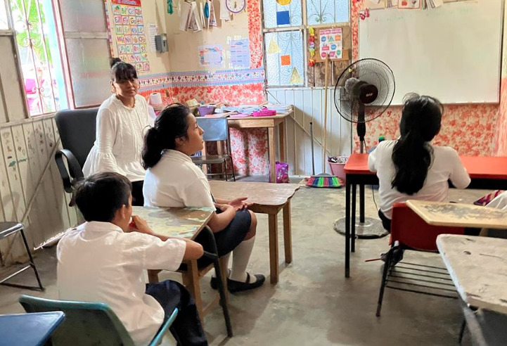 Interior de un salón de clases en Baja California donde estudiantes y una docente permanecen frente a un ventilador de pedestal para mitigar el calor extremo.