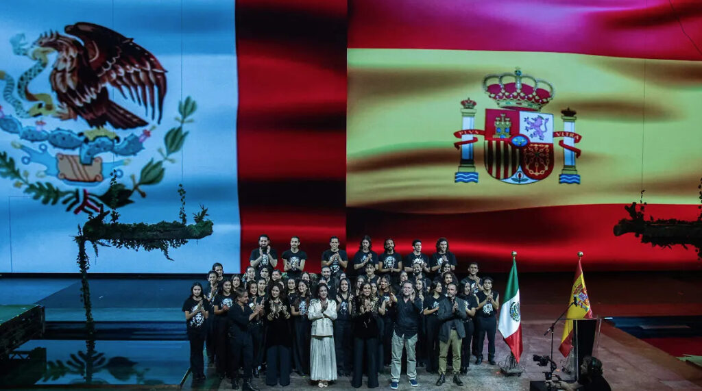 Evento cultural frente a pantallas gigantes con las banderas de México y España, donde se conmemora la evangelización y el mestizaje en México.