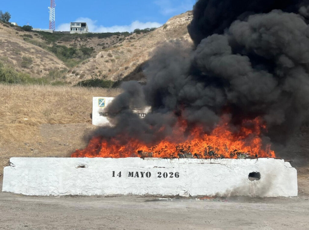 Gran columna de humo negro y fuego durante la incineración de más de siete toneladas de drogas por la FGR en Baja California, con la fecha 14 de mayo de 2026 rotulada en un muro blanco.