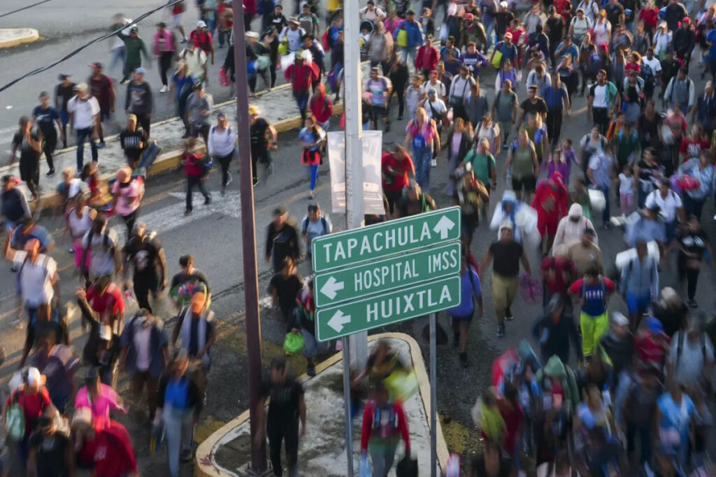 Multitud de personas caminando por una carretera en Tapachula, pasando junto a señalamientos viales que indican las direcciones hacia Tapachula, Hospital IMSS y Huixtla.
