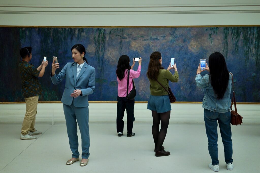 Varios visitantes de un museo de espaldas y de perfil, sosteniendo sus teléfonos celulares para tomar fotos de un enorme cuadro impresionista de nenúfares azules en la película Los Colores del Tiempo.