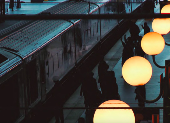 Fotograma cinematográfico de la película 'Geografía humana' que muestra la silueta de un tren en una estación nocturna con lámparas globulares encendidas y sombras de personas en el andén.